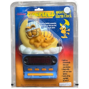 May include: A Sunbeam Garfield nightlight alarm clock in original plastic packaging. The clock features a yellow Garfield character wearing a striped nightcap, resting on a yellow crescent moon and white clouds above a blue digital clock base.