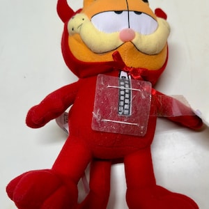 May include: Red plush toy of Garfield dressed as a devil. He has a red suit with a black zipper and a red bow tie. He has a yellow mustache and orange fur.