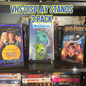 以下が含まれることがあります： 3パックのラベルが付いた黒いVHSディスプレイスタンド3個セット。スタンドには、モンスターズ・インク、ハリー・ポッターと賢者の石、ホーカス・ポカスなど、さまざまな映画のVHSテープが置かれています。