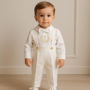 May include: A white baby boy outfit with a gold bow tie and a gold embroidered monogram on the chest. The outfit includes a long-sleeved shirt, trousers, and braces.