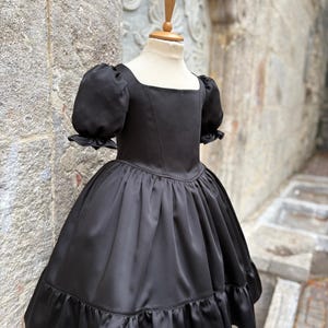 Puede incluir: Un vestido negro liso con escote cuadrado, cuerpo entallado con costuras verticales y mangas cortas abullonadas con bordes con volantes. La falda est&aacute; fruncida en la cintura e incluye un dobladillo ancho con volantes en la parte inferior.