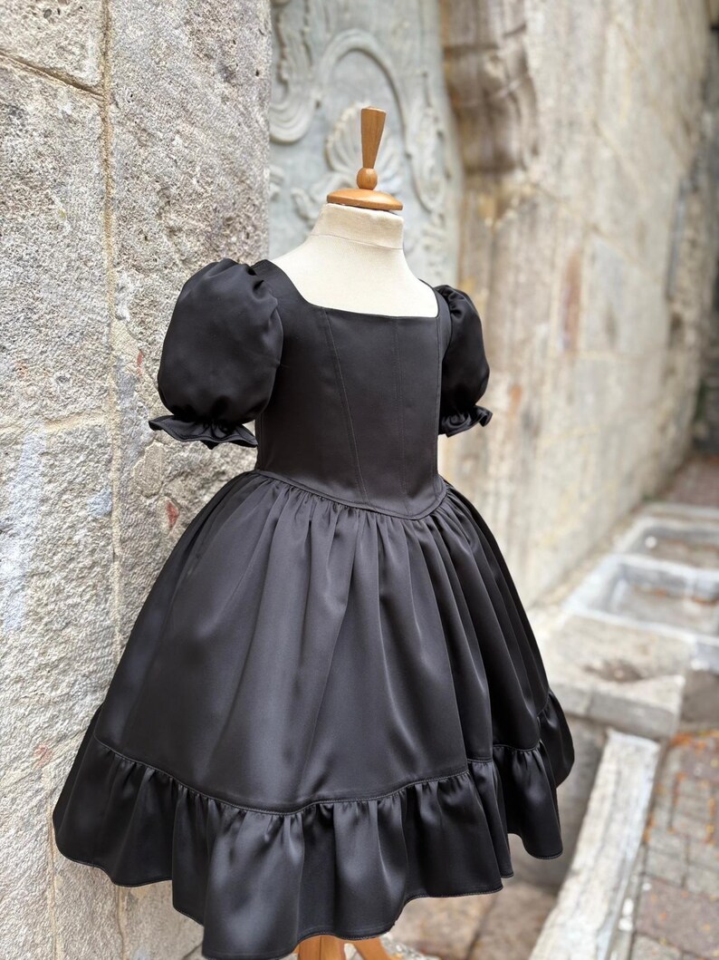 Puede incluir: Un vestido negro liso con escote cuadrado, cuerpo entallado con costuras verticales y mangas cortas abullonadas con bordes con volantes. La falda est&aacute; fruncida en la cintura e incluye un dobladillo ancho con volantes en la parte inferior.