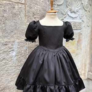 Puede incluir: Un vestido negro liso con escote cuadrado, mangas cortas abullonadas con bordes con volantes, un corpi&ntilde;o ajustado y una falda amplia y fruncida que termina en un dobladillo con volantes. La prenda se exhibe en un torso de maniqu&iacute;.
