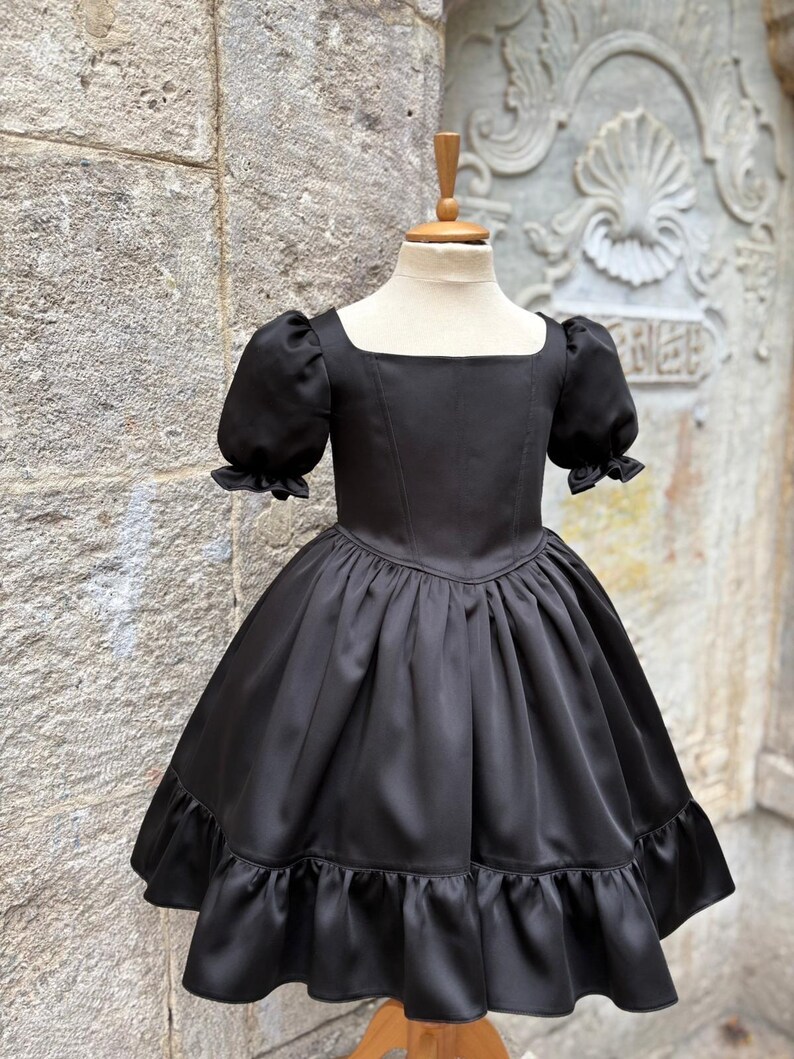 Puede incluir: Un vestido negro liso con escote cuadrado, mangas cortas abullonadas con bordes con volantes, un corpi&ntilde;o ajustado y una falda amplia y fruncida que termina en un dobladillo con volantes. La prenda se exhibe en un torso de maniqu&iacute;.