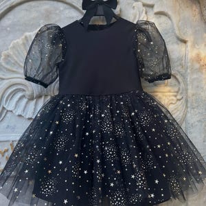 Puede incluir: Un vestido negro con falda de tul y mangas transparentes. La falda y las mangas est&aacute;n adornadas con un patr&oacute;n de estrellas doradas. El vestido est&aacute; colgado de una percha negra con un lazo.