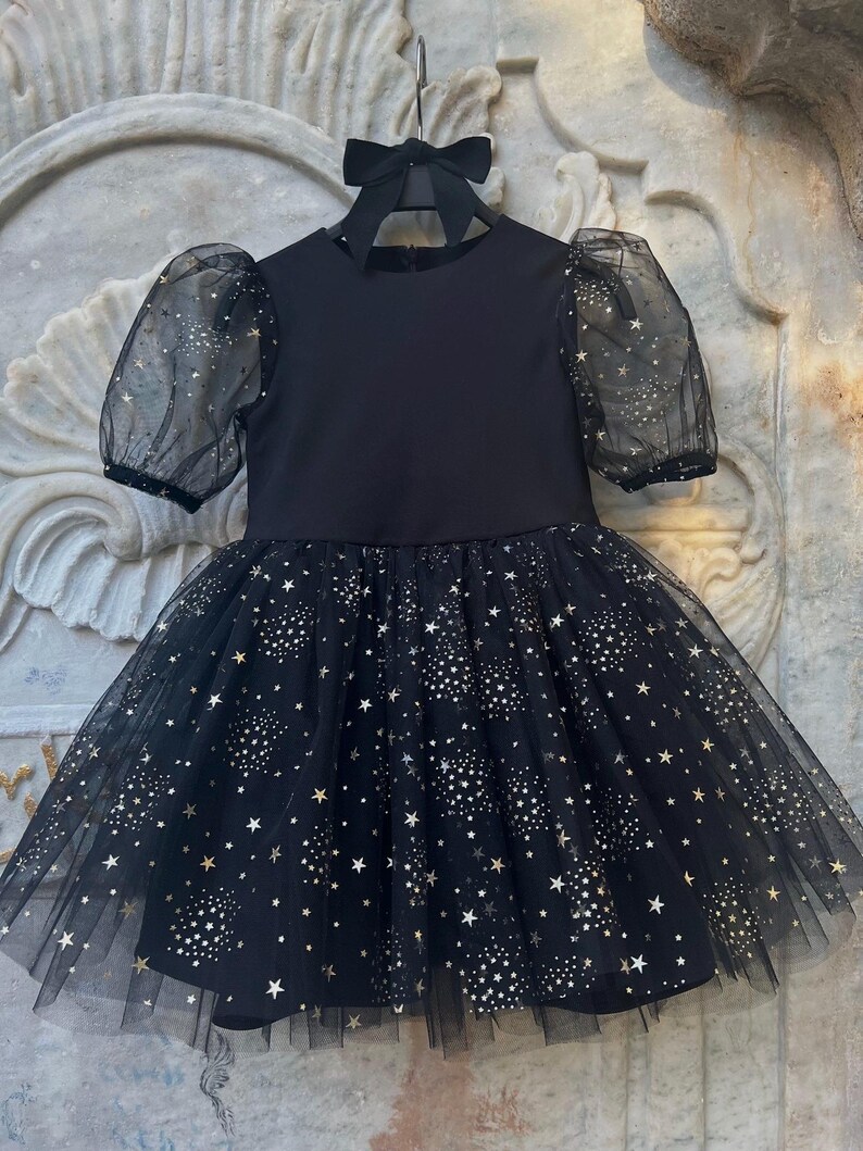 Puede incluir: Un vestido negro con falda de tul y mangas transparentes. La falda y las mangas est&aacute;n adornadas con un patr&oacute;n de estrellas doradas. El vestido est&aacute; colgado de una percha negra con un lazo.