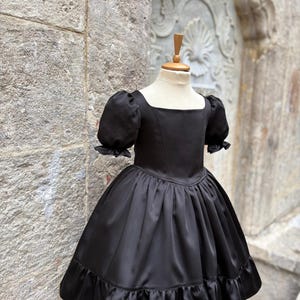 Puede incluir: Un vestido negro con escote cuadrado, mangas cortas abullonadas con bordes con volantes y un corpi&ntilde;o ajustado. La falda est&aacute; fruncida en la cintura, con una silueta completa y un dobladillo ancho con volantes, exhibido en un maniqu&iacute;.