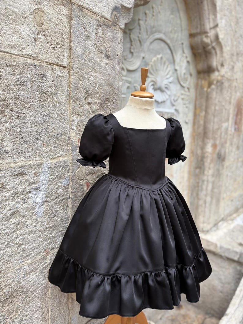 Puede incluir: Un vestido negro con escote cuadrado, mangas cortas abullonadas con bordes con volantes y un corpi&ntilde;o ajustado. La falda est&aacute; fruncida en la cintura, con una silueta completa y un dobladillo ancho con volantes, exhibido en un maniqu&iacute;.