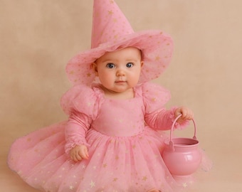 May include: A pink dress with puff sleeves and a matching pointed hat, both featuring a gold star pattern. The dress has a full, layered tulle skirt. A small, solid pink plastic bucket with a handle is held in one hand, completing the ensemble.