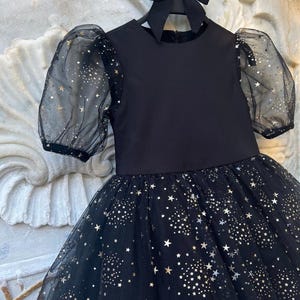 Puede incluir: Un vestido negro con falda de tul y mangas transparentes, adornado con un estampado de estrellas doradas. El vestido tiene un cuello redondo y un lazo negro a juego. El vestido est&aacute; colgado de una percha negra.