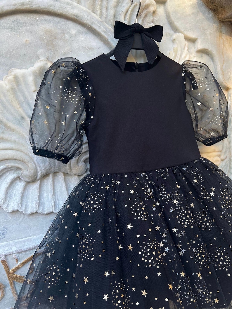 Puede incluir: Un vestido negro con falda de tul y mangas transparentes, adornado con un estampado de estrellas doradas. El vestido tiene un cuello redondo y un lazo negro a juego. El vestido est&aacute; colgado de una percha negra.