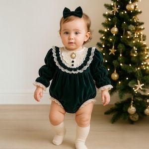 May include: A baby wearing a dark green velvet romper with a white lace collar and cuffs, and a matching bow. The romper has a decorative gold button. The baby is wearing white socks and standing in front of a Christmas tree.