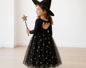 May include: A black witch costume for children, featuring a long-sleeved leotard with a star-patterned tulle skirt. The outfit includes a pointed witch hat and a star-shaped wand. The back of the leotard has a bow detail. The child is wearing black shoes.