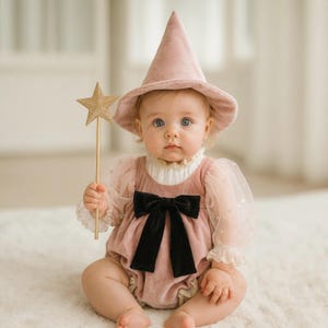 Puede incluir: Un sombrero puntiagudo de terciopelo rosa empolvado y un pelele de terciopelo a juego con mangas abullonadas transparentes, un cuello alto con volantes y un gran lazo de terciopelo negro en el centro. Se sostiene una varita de estrella dorada.