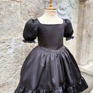 Puede incluir: Un vestido negro liso con escote cuadrado, cuerpo entallado con costuras verticales y mangas cortas abullonadas con bordes con volantes. La falda est&aacute; fruncida en la cintura, creando una silueta amplia, y termina con un dobladillo con volantes.