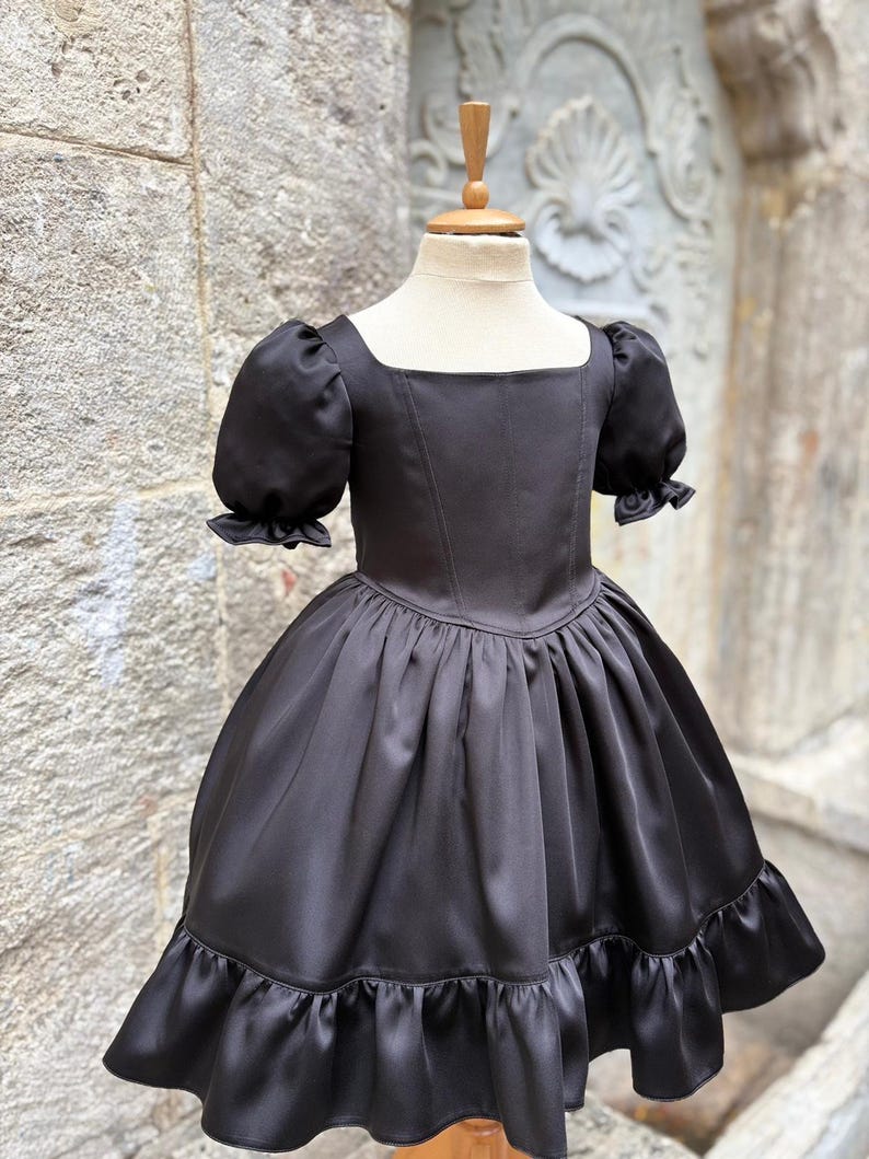 Puede incluir: Un vestido negro liso con escote cuadrado, cuerpo entallado con costuras verticales y mangas cortas abullonadas con bordes con volantes. La falda est&aacute; fruncida en la cintura, creando una silueta amplia, y termina con un dobladillo con volantes.