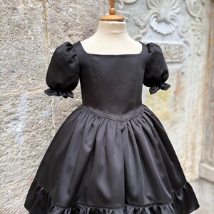 Puede incluir: Un vestido negro con escote cuadrado, mangas cortas abullonadas con pu&ntilde;os con volantes y un corpi&ntilde;o ajustado. La falda est&aacute; fruncida en la cintura, creando una silueta amplia, y termina con un dobladillo ancho con volantes en el borde.