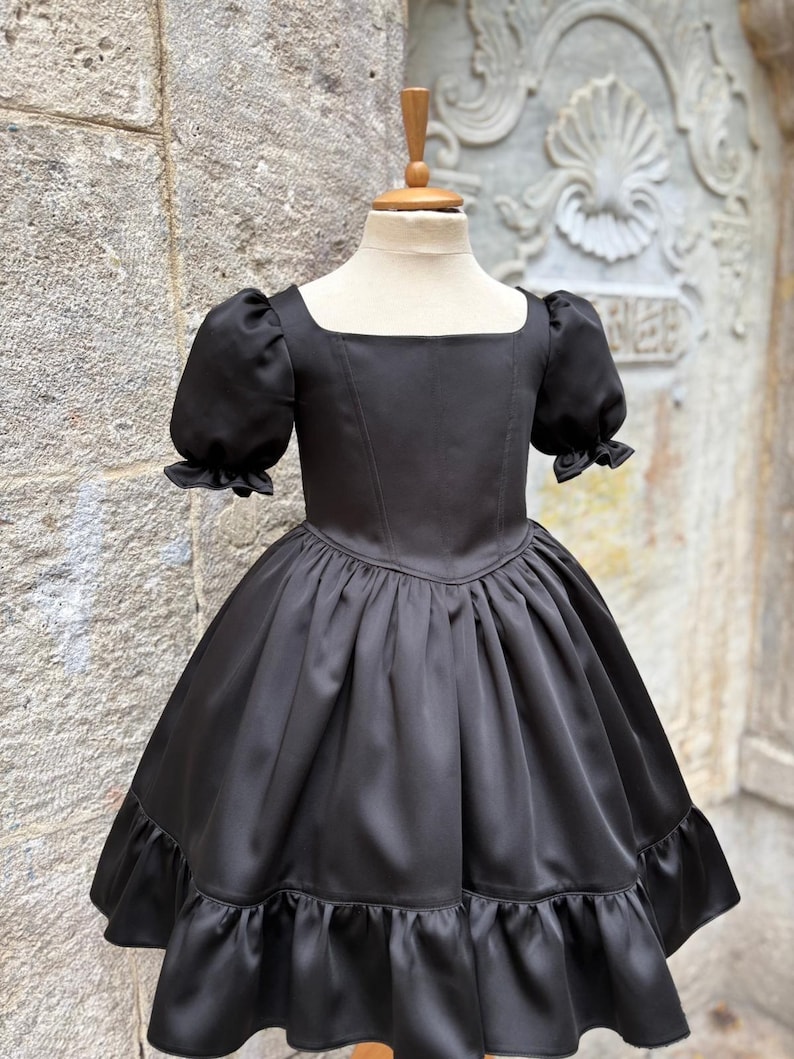 Puede incluir: Un vestido negro con escote cuadrado, mangas cortas abullonadas con pu&ntilde;os con volantes y un corpi&ntilde;o ajustado. La falda est&aacute; fruncida en la cintura, creando una silueta amplia, y termina con un dobladillo ancho con volantes en el borde.