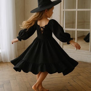 Puede incluir: Un disfraz de bruja negro para niños, que incluye un vestido de manga larga con dobladillo con volantes y un sombrero puntiagudo a juego. El vestido tiene un escote cuadrado y un detalle de espalda con cordones. El niño está girando sobre un suelo de madera.