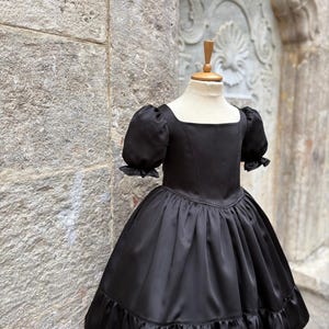 Puede incluir: Un vestido negro liso con escote cuadrado, mangas cortas abullonadas con pu&ntilde;os de volantes, un corpi&ntilde;o ajustado y una falda amplia fruncida con un dobladillo ancho de volantes. La prenda se exhibe sobre un maniqu&iacute;, mostrando su textura suave.
