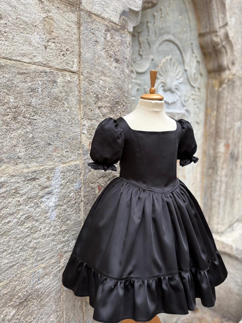 Puede incluir: Un vestido negro liso con escote cuadrado, mangas cortas abullonadas con pu&ntilde;os de volantes, un corpi&ntilde;o ajustado y una falda amplia fruncida con un dobladillo ancho de volantes. La prenda se exhibe sobre un maniqu&iacute;, mostrando su textura suave.