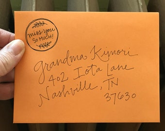 May include: An orange envelope with handwritten address details and a circular stamp that reads "miss you so much!". The address is for Grandma Kimori, 402 Iota Lane, Nashville, TN 37630. The stamp is surrounded by a decorative leaf design.