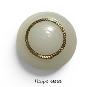 Può includere: Un bottone rotondo in vetro bianco sporco con una finitura dorata a forma di corda. Il bottone ha una forma leggermente bombata e una superficie liscia. Il testo "Hoppe Glass" è stampato in basso nell'immagine.