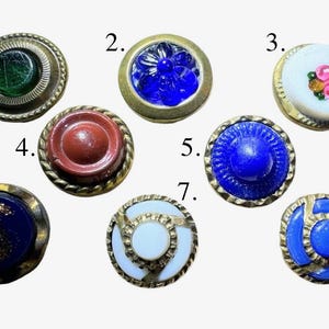 May include: A collection of eight vintage buttons in various colors and designs. The buttons feature a range of materials, including glass and metal, with decorative elements like floral patterns and textured surfaces. The buttons are round and vary in size.