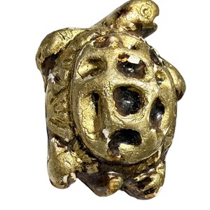 May include: A small, gold-colored turtle figurine with a textured, aged appearance. The turtle's shell features a unique pattern of holes and dark spots, and the figurine has a weathered, antique look.