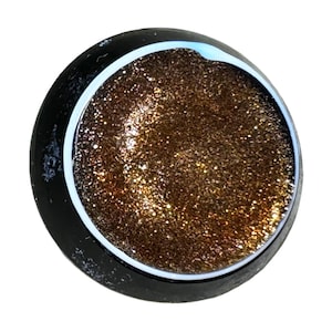 Può includere: Un oggetto rotondo nero con un anello interno bianco e un centro dorato scintillante e glitterato. I glitter dorati sono fittamente impacchettati, creando un effetto scintillante. L'oggetto è posto su uno sfondo bianco.