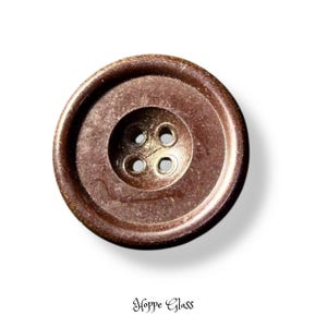 May include: A round, dark brown button with a smooth, slightly metallic finish and four central holes. The button features a raised outer rim and a recessed center. Below the button, the text Hoppe Glass is printed in a black script font.