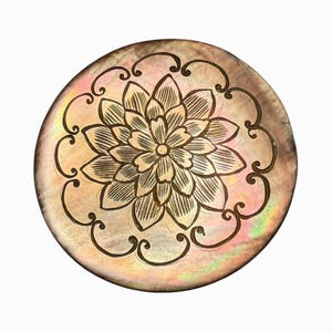 May include: Round, iridescent mother-of-pearl decorative piece with a detailed floral design in the center. The flower is surrounded by an ornate, etched border. The piece has a pearlescent sheen with hints of pink, gold, and green.