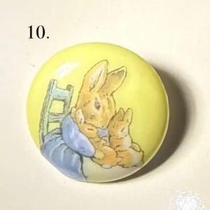 Puede incluir: Un botón redondo y amarillo con una ilustración en acuarela de una madre coneja y dos conejitos. La madre coneja lleva un vestido azul y está sentada en una silla. El botón tiene el número 10 en la esquina superior izquierda.