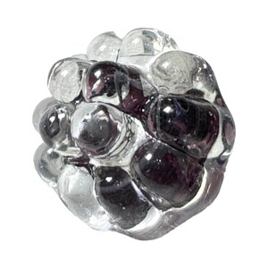 Può includere: Una scultura in vetro trasparente che raffigura un grappolo d'uva. La scultura presenta una miscela di sfere di vetro trasparente e viola scuro, creando una forma strutturata e organica. Il pezzo è posto su uno sfondo bianco.