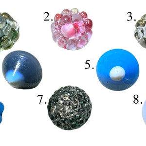 Può includere: Una collezione di otto oggetti decorativi in vetro di varie forme e colori. Gli oggetti includono sfere, grappoli e forme ovali, con colori che vanno dal trasparente e verde al blu e rosa. Alcuni hanno superfici strutturate.