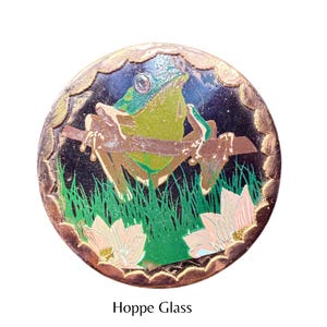 Könnte beinhalten: Runder Glasanhänger mit einem Frosch auf einem Ast, umgeben von Gras und Blumen. Der Frosch ist grün und gelb, vor dunklem Hintergrund. Mit goldfarbenem, gewelltem Rand und dem Text "Hoppe Glass".