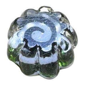 Può includere: Una perla di vetro trasparente a forma di fiore. La perla presenta un motivo a spirale argentata al centro e strisce bianche sui bordi. Il vetro ha sfumature verdi e grigie.