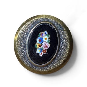 May include: A round brooch featuring a textured brass-toned metal border surrounding a central oval black inlay. The black center is decorated with a small, colorful micro-mosaic floral arrangement consisting of blue, yellow, red, and white petals.