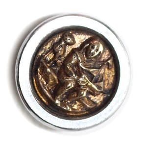 May include: A circular button featuring a polished silver-toned outer rim and a recessed, textured bronze-colored center. The center displays a raised, detailed relief of a figure in a hooded garment, appearing to be engaged in a manual task.