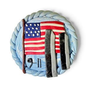 May include: A circular pin featuring a light blue braided border and a central design of the American flag. Two vertical black rectangular shapes represent towers, and the text 9-11 is inscribed in black at the bottom left of the blue background.
