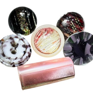 May include: A collection of decorative glass buttons and beads in various shapes and colors. Includes a black and gold striped bead, a white button with purple speckles, and a pink rectangular bead. The items are arranged on a white background.