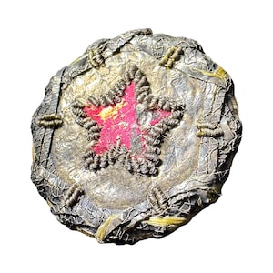 May include: A circular decorative item with a central red star. The star is outlined with silver wire, and the outer edge has a woven, textured design. Gold and yellow accents are visible on the metallic surface.