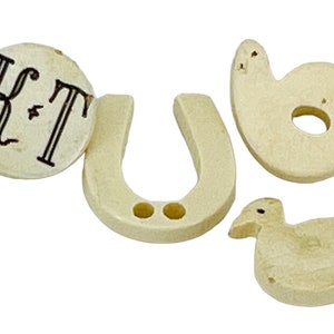 May include: A collection of off-white, carved wooden shapes. Includes a round piece with "K+T" in black script, a horseshoe, a number six, and a duck. The objects are likely decorative or for crafts.