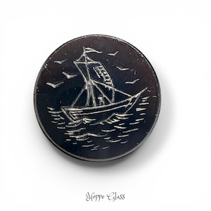 May include: A dark, circular glass button with a detailed etched design of a sailboat on the water, with birds in the sky. The button has a shiny, reflective surface. The text "Hoppe Glass" is at the bottom.