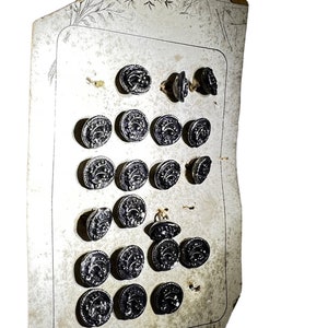 May include: A collection of nineteen circular dark metallic buttons with embossed floral patterns, arranged in rows on a rectangular off-white paper card featuring a faint decorative line drawing and a border at the top edge.