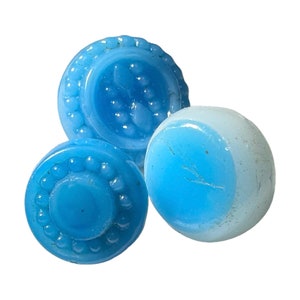 Può includere: Tre biglie e bottoni antichi in vetro blu. Due bottoni hanno un design circolare con un bordo perlato. La biglia è di colore azzurro e bianco. Gli oggetti sono su uno sfondo bianco.