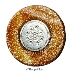 Può includere: Un bottone rotondo e decorativo con un anello esterno dorato glitterato e un centro argentato. Il centro presenta un motivo floreale in rilievo. Il testo "KPHoppeGlass" è in basso.
