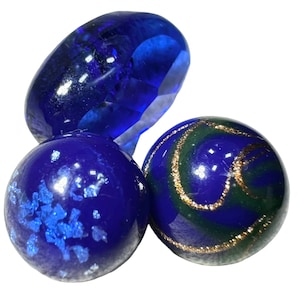 Può includere: Tre biglie decorative in vetro blu. Una è ovale e sfaccettata, le altre sono rotonde. Una biglia rotonda ha un vortice di glitter verdi e dorati. L'altra biglia rotonda ha glitter blu.