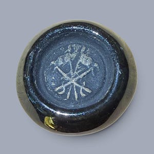May include: A circular, dark-colored ceramic object with a glossy finish. The center features an embossed, light-colored stamp depicting two crossed tools or implements, possibly representing a crest or maker's mark, set within a recessed area.