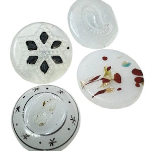 Può includere: Quattro bottoni decorativi trasparenti con design unici. Un bottone presenta un design a fiocco di neve con forme a diamante nere. Un altro ha un motivo a paisley. Il terzo ha accenti rossi e dorati, e l'ultimo ha un design a stella e mezzaluna nera.